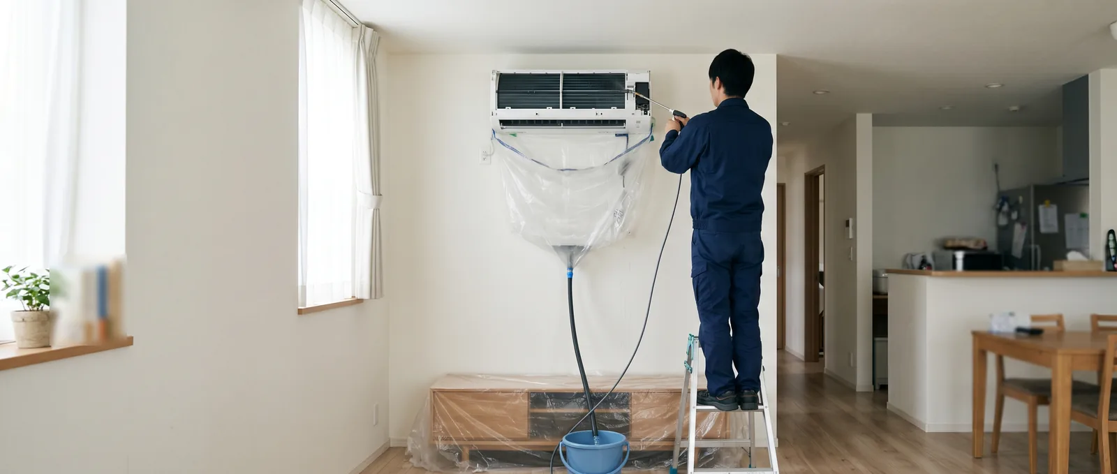 リビングの壁掛けエアコンに洗浄カバーを付け、脚立に乗った作業者が水で洗っている様子